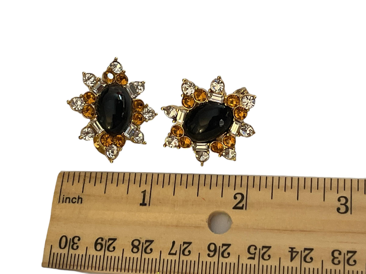 A pair of statement earrings with a black stone center surrounded by a floral design and small diamonds, placed next to a ruler for size comparison.
