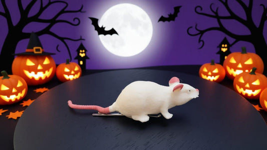 A white toy rat sits on a black surface, surrounded by carved jack-o-lanterns and bats against a purple and black night sky with a full moon.