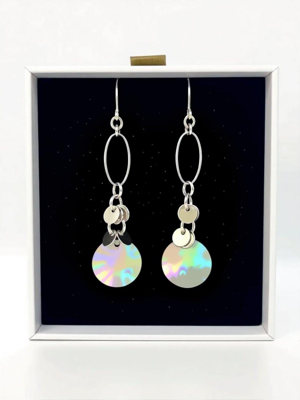 A pair of silver earrings with circular designs hanging from the ear hooks, displayed in a white box against a black background.