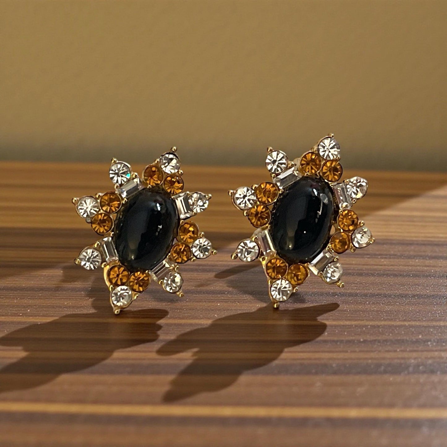 A pair of elegant earrings with a black gemstone surrounded by sparkling rhinestones and gold accents, resting on a wooden surface.