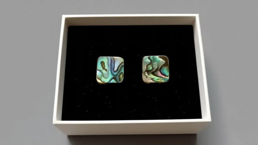 A hand holding two square earrings with a green and blue pattern, placed in front of a white box.