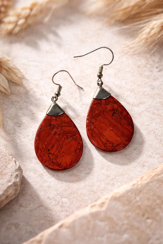 A pair of red, oval-shaped earrings with a metal hook and a small, round metal piece at the top, resting on a beige surface.