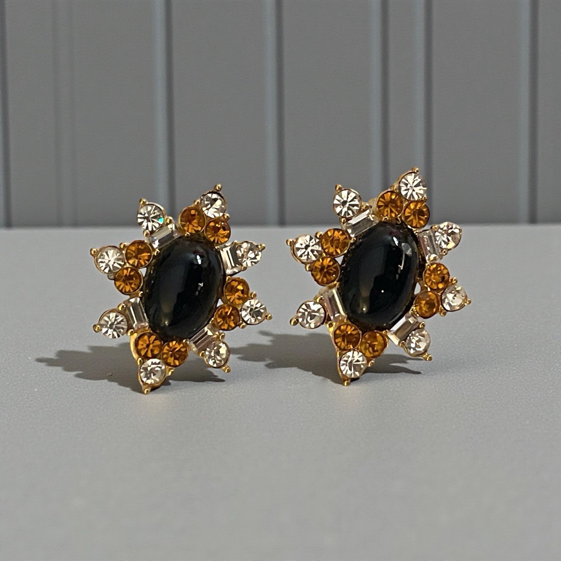 A pair of elegant earrings with a black gemstone surrounded by gold and white gemstones, set against a gray background.