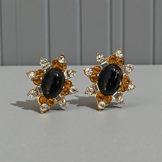 A pair of elegant earrings with a black gemstone surrounded by gold and white gemstones, set against a gray background.