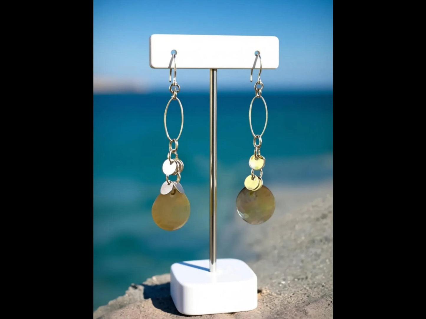A pair of earrings with circular charms hanging from them, displayed on a white stand against a backdrop of a blue sky and water.