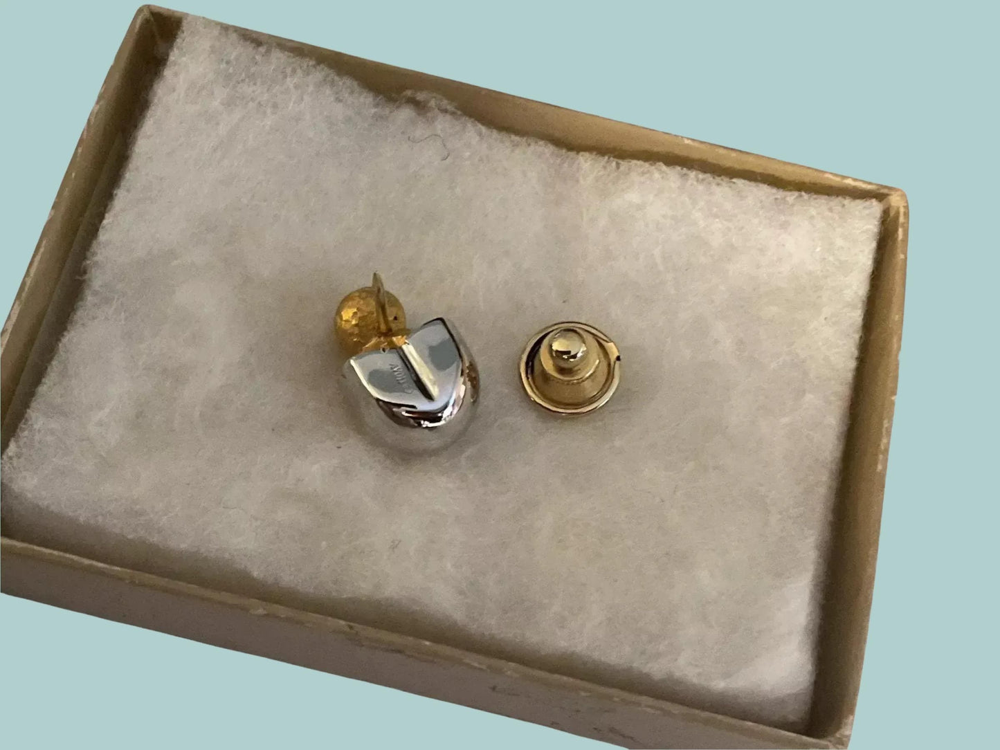 A small, silver-colored object with a gold-colored clasp is displayed in a box, which is placed on a white cloth.