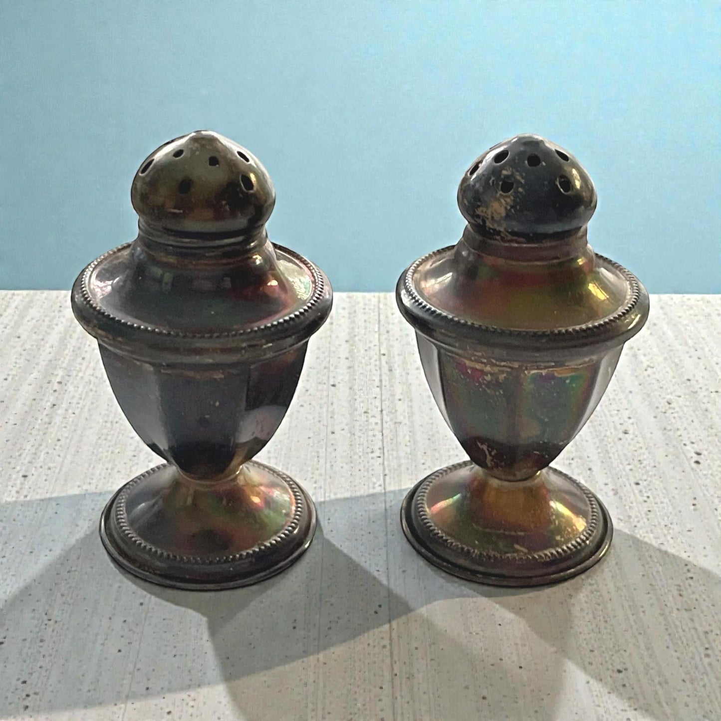 Two antique-looking salt and pepper shakers with a metallic finish are placed side by side on a wooden surface against a blue background.