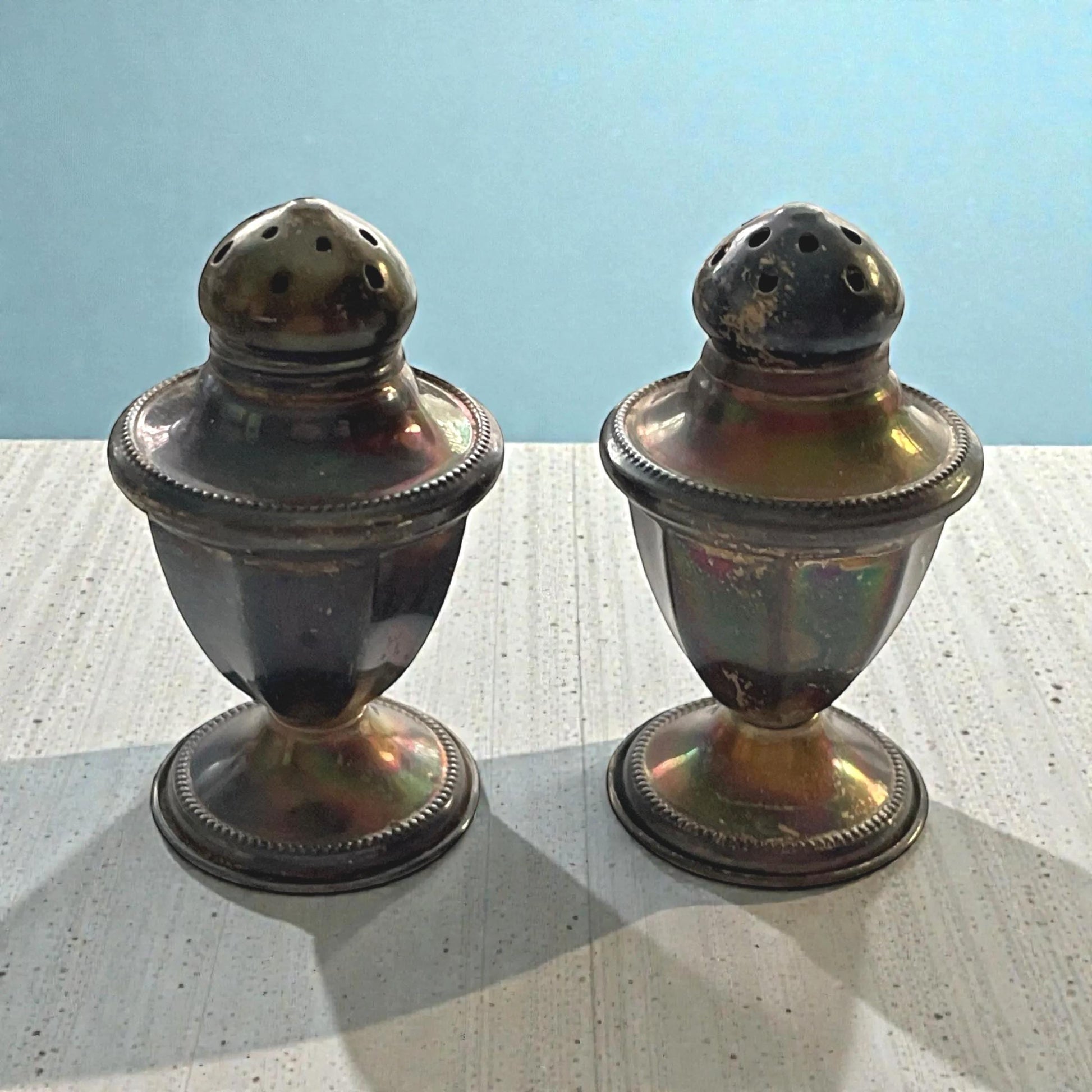 Two antique-looking salt and pepper shakers with a metallic finish are placed side by side on a wooden surface against a blue background.