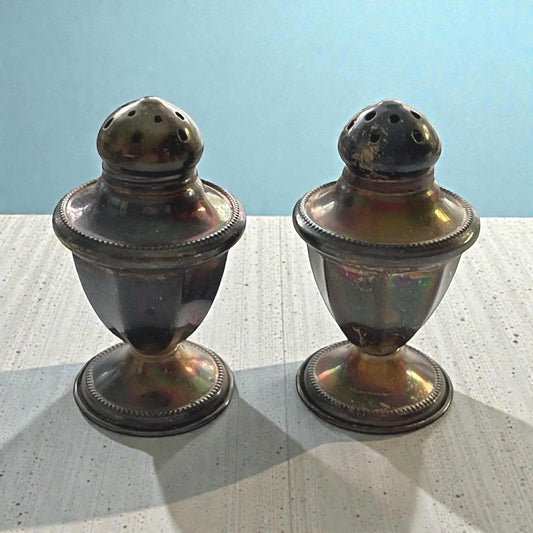 Two antique-looking salt and pepper shakers with a metallic finish are placed side by side on a wooden surface against a blue background.